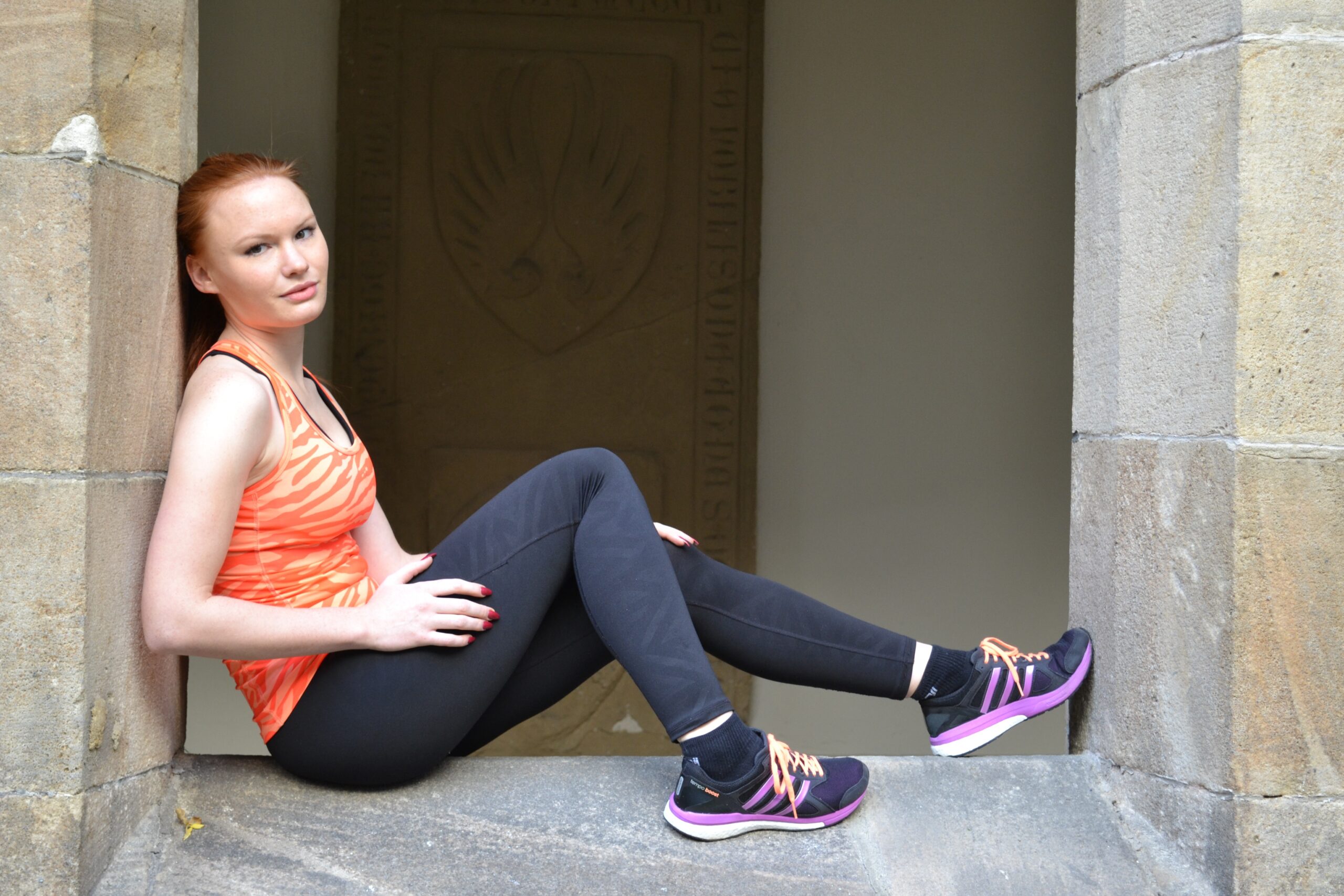 Woman in an orange athletic top and black leggings sits in a stone window alcove, legs extended, wearing purple sneakers.
