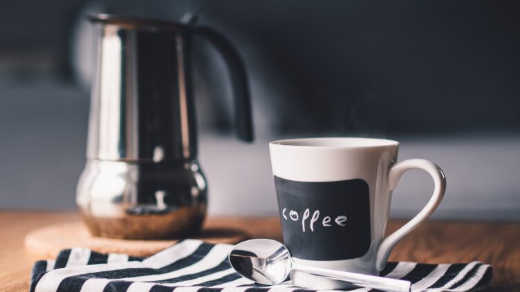 White coffee cup with 'coffee' text on it, placed on a striped napkin with a blurred metal moka pot in the background.