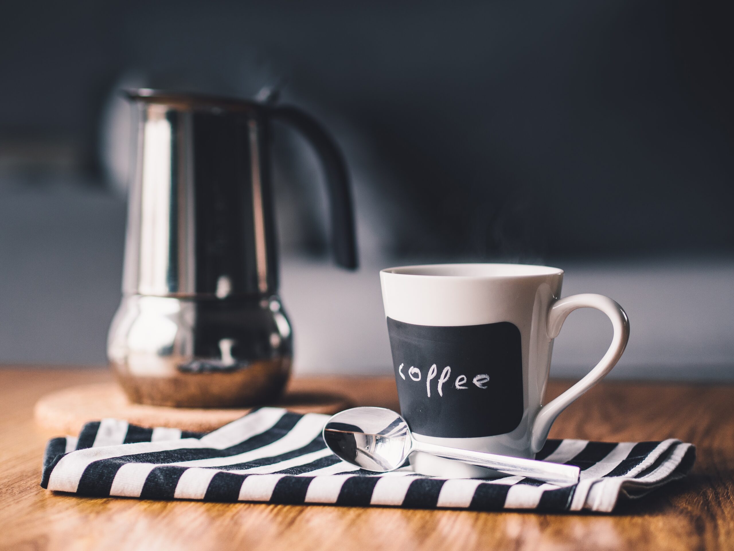 White coffee cup with 'coffee' text on it, placed on a striped napkin with a blurred metal moka pot in the background.