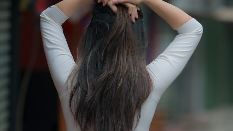 Person viewed from behind with long hair gathered at the nape, hands clasped on top of the head wearing a white long-sleeve top