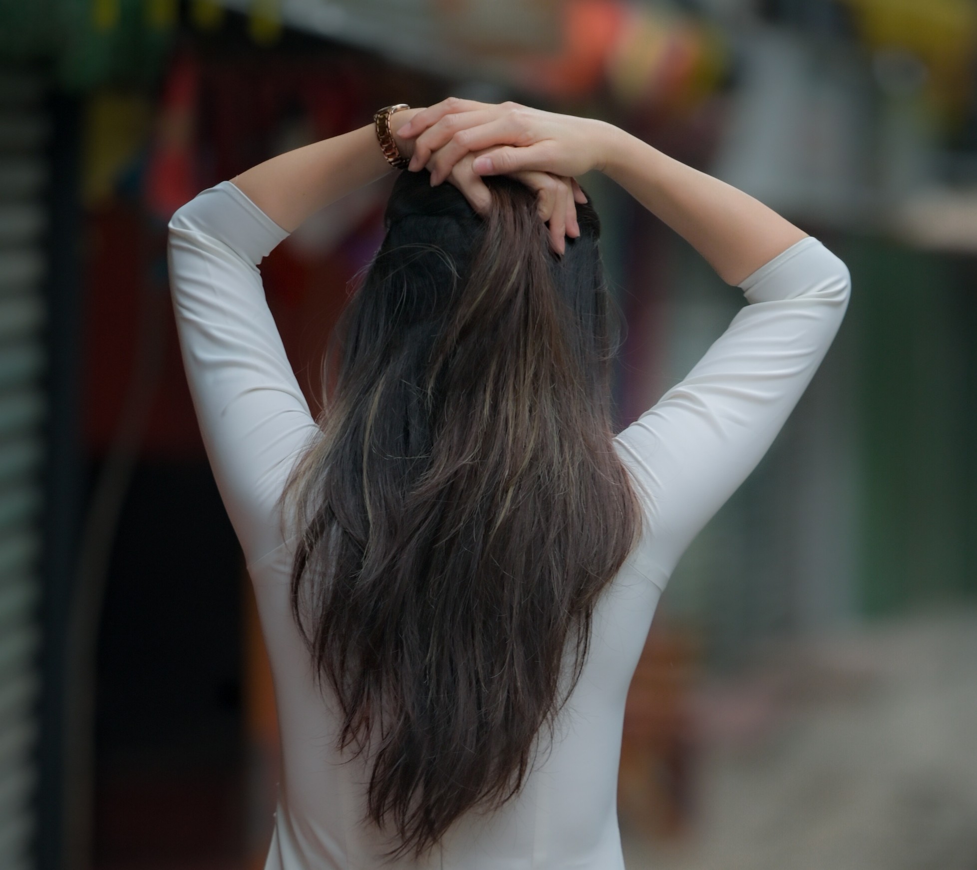 Person viewed from behind with long hair gathered at the nape, hands clasped on top of the head wearing a white long-sleeve top