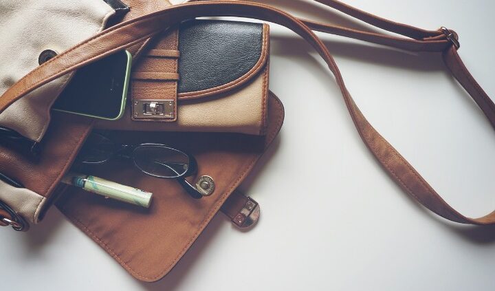 Brown leather handbag with a phone, glasses, wallet, and a pen visible in an open pocket area on a light background.