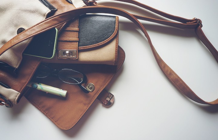 Brown leather handbag with a phone, glasses, wallet, and a pen visible in an open pocket area on a light background.