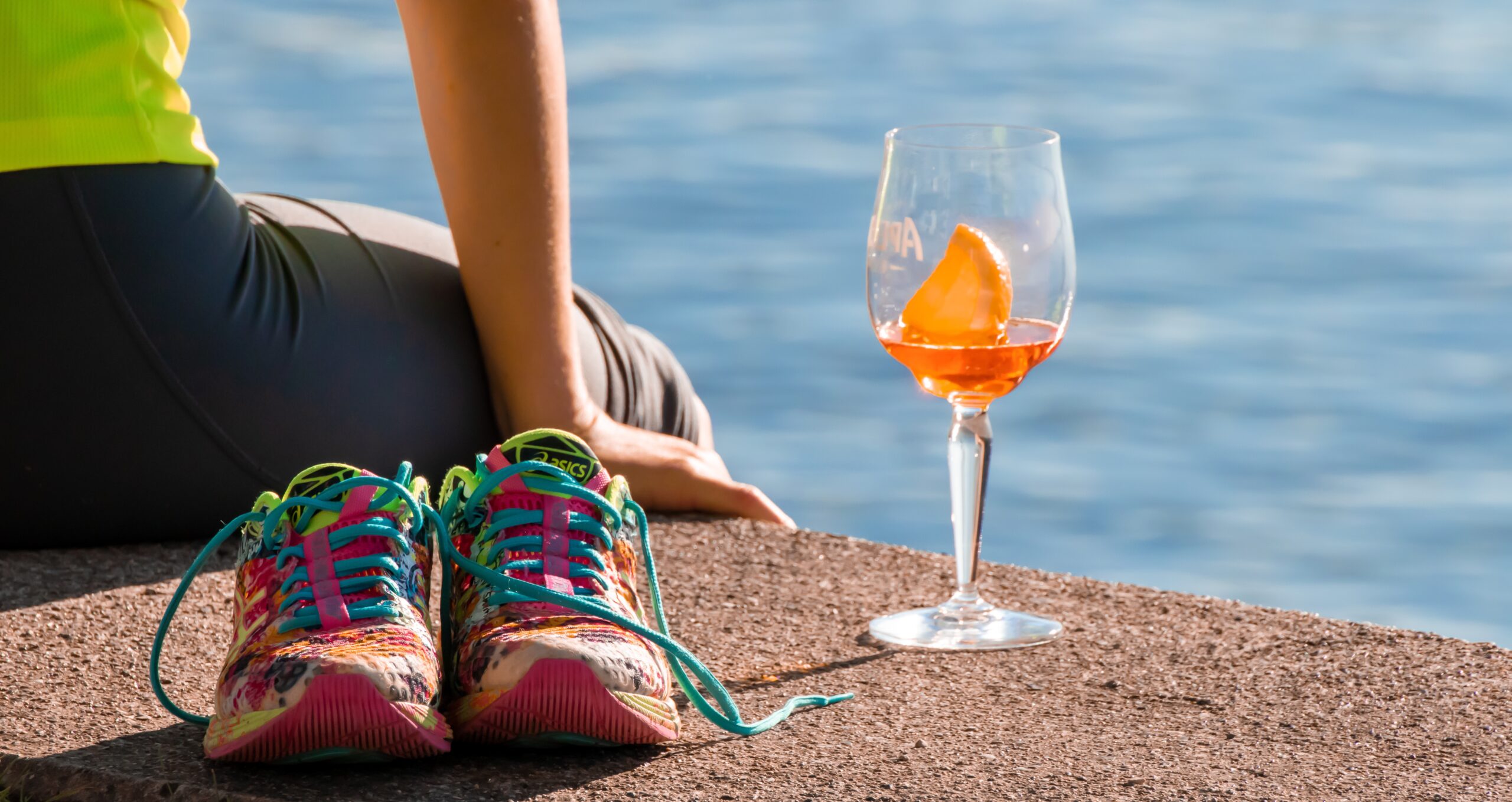 Person in a neon shirt sits by the water with colorful sneakers and an orange cocktail glass nearby.