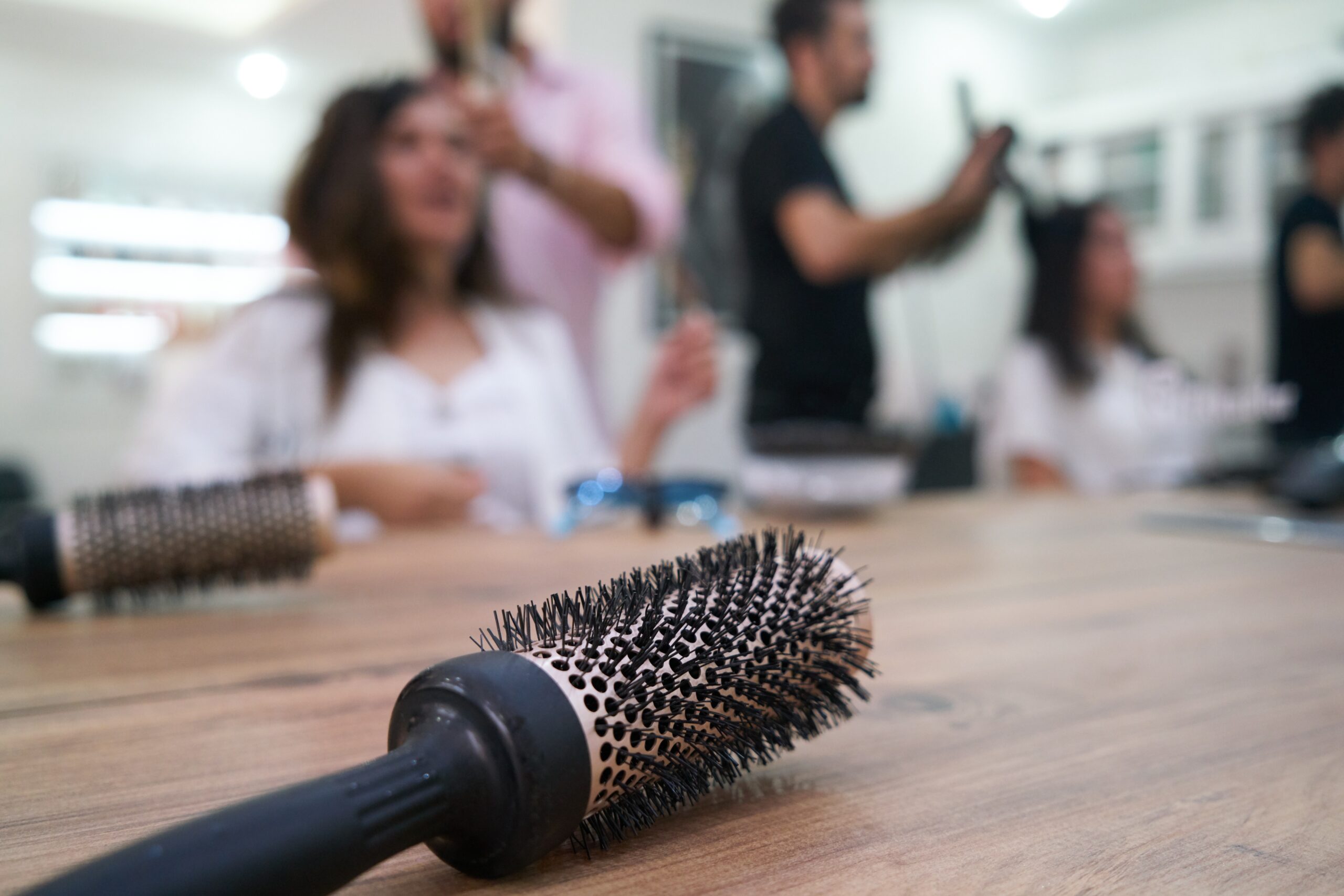 Close-up of a round hairbrush on a wooden table with blurred hairstylists and clients in a salon background.
