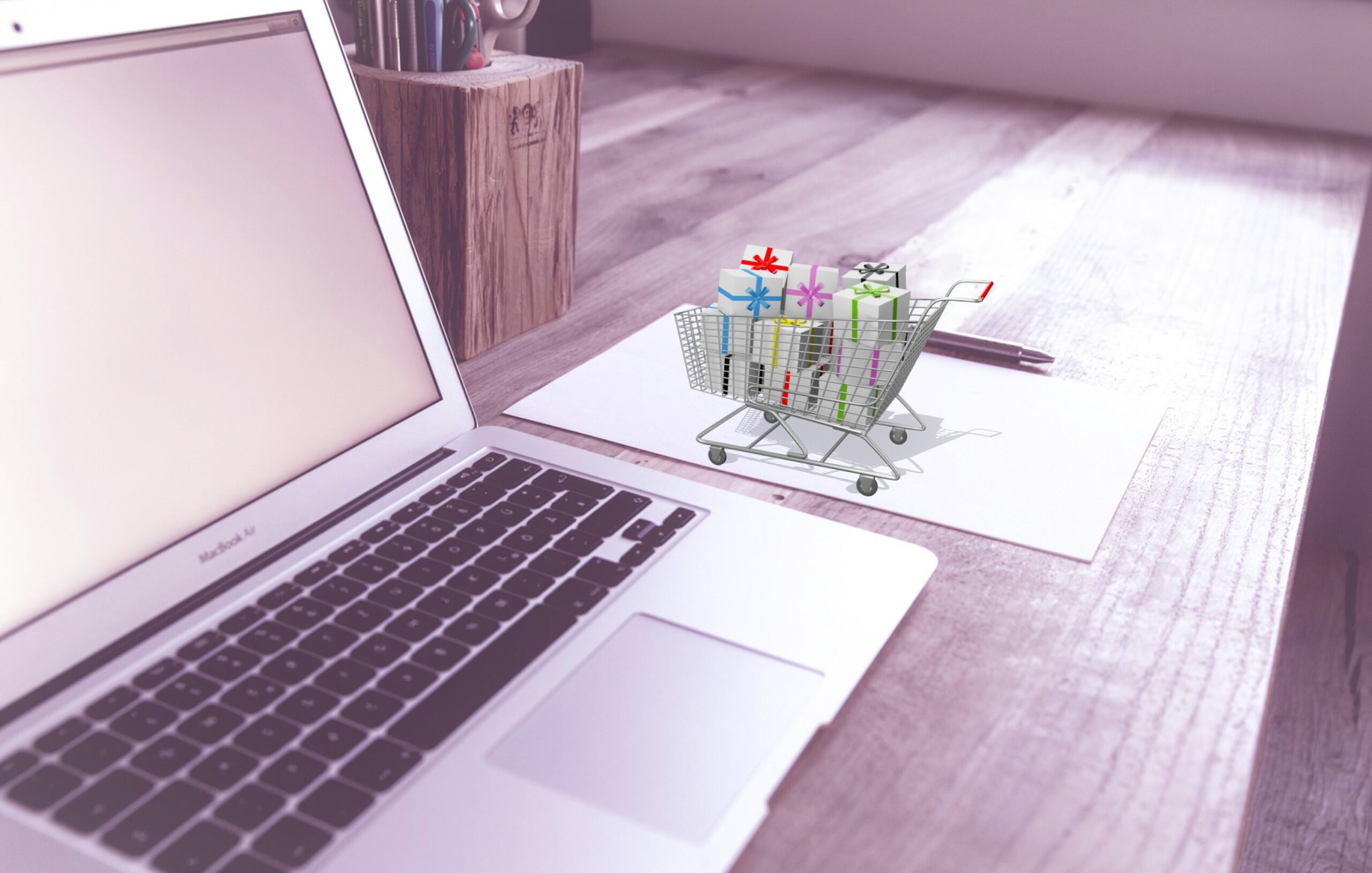Laptop on a desk with a small shopping cart filled with wrapped gift boxes, suggesting online shopping for gifts.