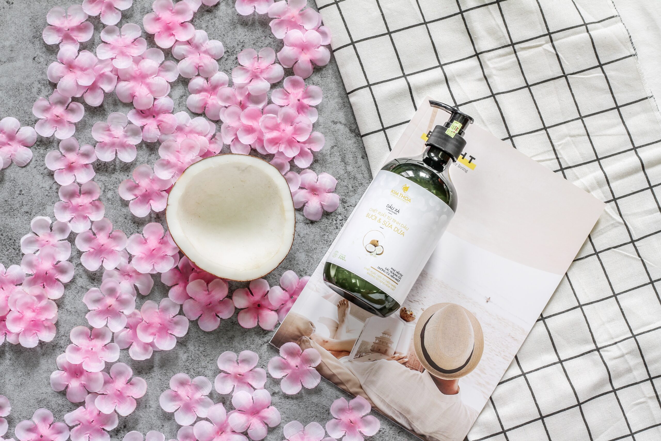Flat lay of pink petal blossoms scattered on a gray surface with half coconut, a pump bottle resting on a magazine, and a black-and-white grid cloth nearby.